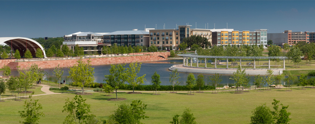 Panoramic photograph of a lake and park with residential and nonresidential buildings in the background.