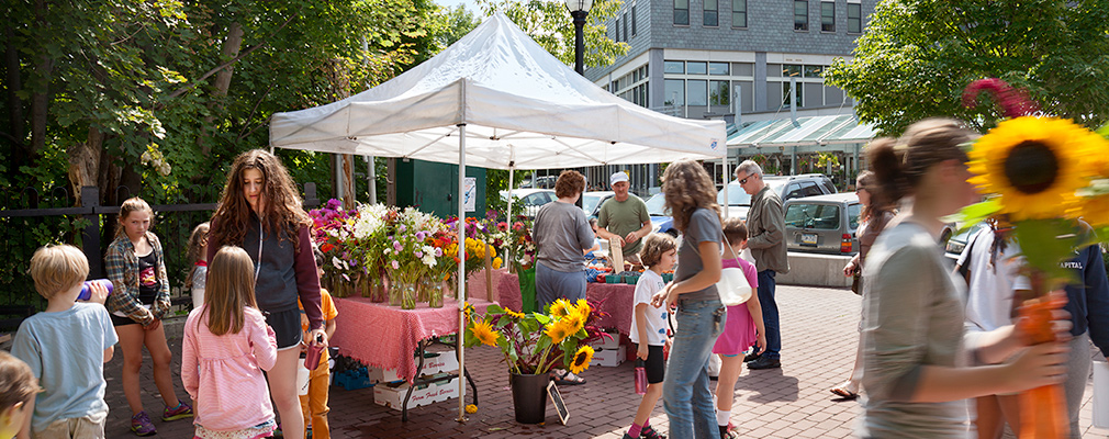 Photograph of a dozen people standing around a flower stand in a plaza.