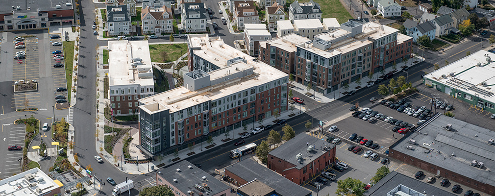 Low-angle aerial photograph showing the development — two 5-story mixed-use buildings, two 4-story apartment buildings, and several town homes — in the context of primarily 2-story commercial buildings and residences.