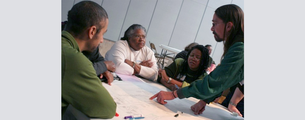Photograph of five people discussing plans laid out on a table inside a large meeting room.