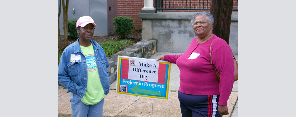 Photograph of two adults standing beside a sign reading “Make A Difference Day. Project in Progress.”