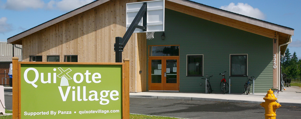 Photograph of a large one-story building with vinyl siding and wood paneling standing behind a sign that reads “Quixote Village: Supported By Panza · quixotevillage.com.”
