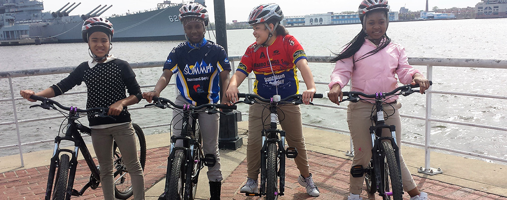 Photograph of four children standing with their bicycles on the Camden Waterfront, with the battleship New Jersey in the background.