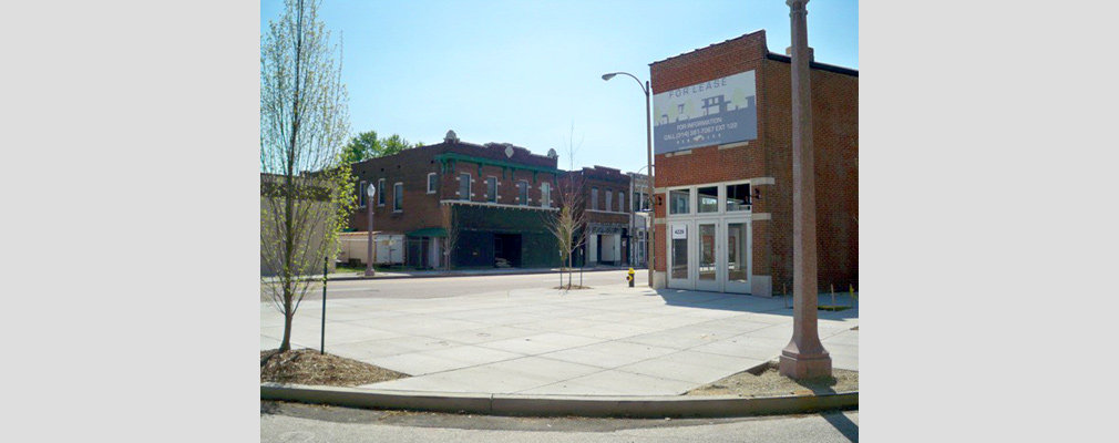 Photograph of a paved plaza with newly planted trees in front of a two-story brick commercial building, with similar buildings in the background across the street.