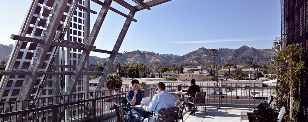 Photograph of three men sitting at tables on rooftop terrace shaded by an array of solar panels.