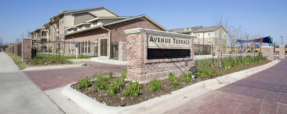 Photograph of the entrance to Avenue Terrace, with a brick monument sign in the street median in the foreground, a one-story community building and playground in the middleground, and three-story multifamily buildings in the background.