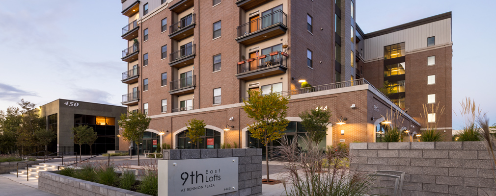 Photograph taken at dusk of the front and side façades of a six-story building that has apartments above retail space on the ground floor, with in the foreground, a sidewalk, a sign reading “9th East Lofts at Bennion Plaza,” and a landscaped plaza. 