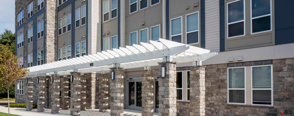 Photograph of the entrance to a four-story multifamily building, accented by a pergola extending across 12 bays of the façade.