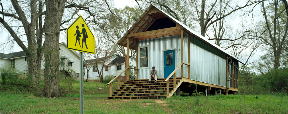 Photograph of a person sitting on the front steps of a one-story single-family house, with two older houses in the background.