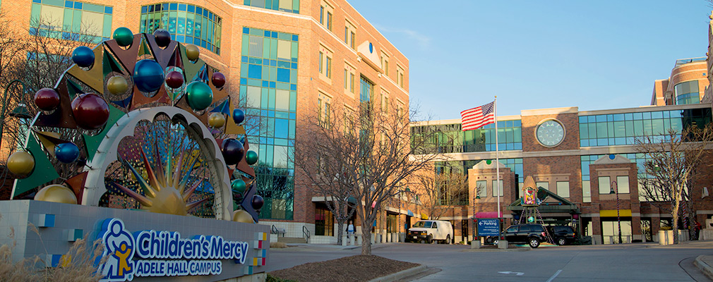 Photograph of a multistory medical building with a sign in the foreground reading "Children’s Mercy Adele Hall Campus."