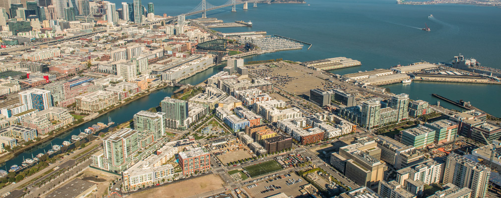 Low-angle aerial photograph of a waterfront neighborhood in San Francisco, with the Financial District and Oakland Bay Bridge in the background. 