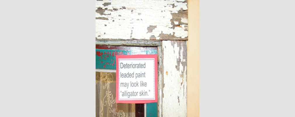 Photograph of chipping paint on a window frame with a sign that reads “Deteriorated leaded paint may look like ‘alligator skin.’”