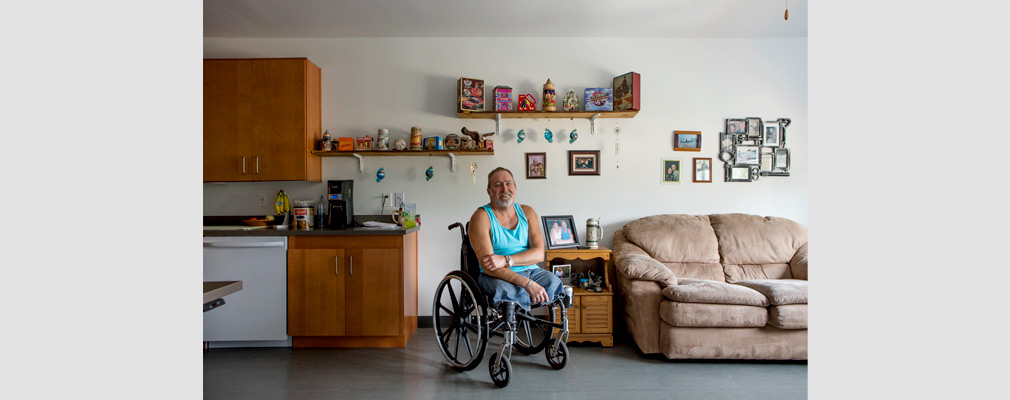 Photograph of a man in a wheelchair in the kitchen-living area of an apartment with various decorations and personal items adorning the wall.