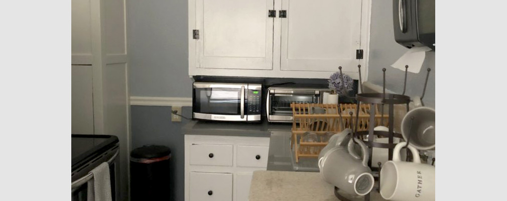 Photograph of a kitchen, with cups, a sink, a microwave, and a toaster oven on a counter between upper and lower cabinets.