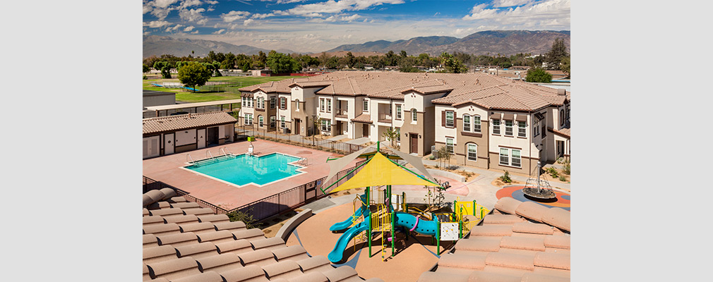 Low-angle aerial photograph of a courtyard with a swimming pool and children’s play structure and a two-story multifamily building in the middle ground.