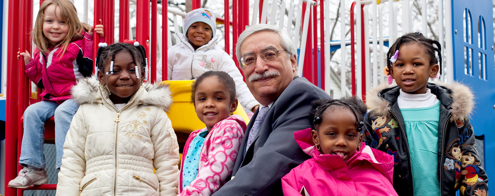 Photograph of six children surrounding a man seated on a piece of playground equipment.