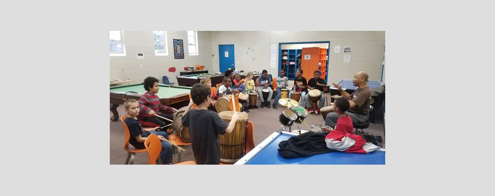 Photograph of a large room with a group of children playing drums and seated in a semicircle  facing an adult instructor.