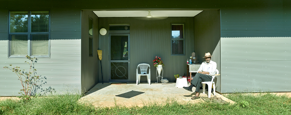 Photograph of a man seated on the front porch of a single-story house that rests on a concrete slab.