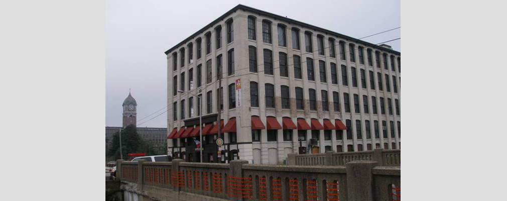 Photograph of two façades of a five-story brick building painted gray, with a canal bridge in the foreground and the tower of an adjacent mill building in the background.