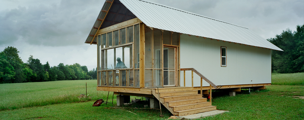 Photograph of a one-story single-family house at the edge of a farm field, with a person standing on a screened porch.