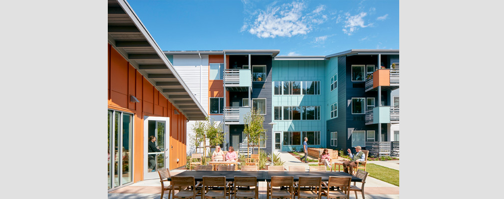 Photograph of a patio with a long table in the foreground, several people sitting and walking in the middle ground, and a three-story residential building in the background. 