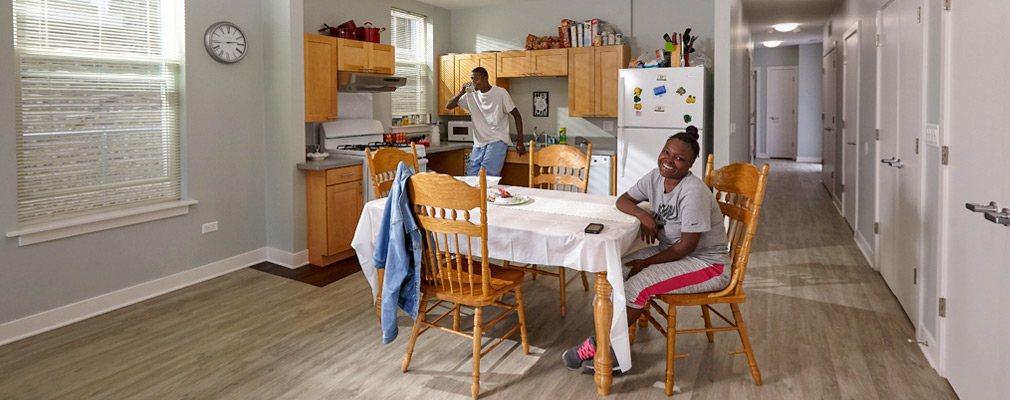 Photograph of two people in an apartment with a large kitchen and dining area and with a wide hallway leading to the rear of the apartment.