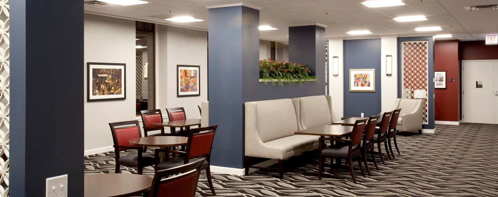 Photograph of a carpeted community lounge with tables, chairs, and upholstered bench seating.