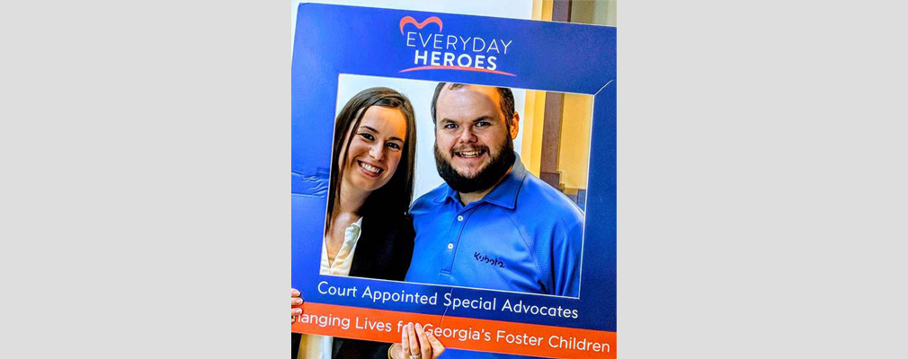 Photograph of two smiling people posing behind a cardboard frame printed with the text: “Everyday Heroes,” “Court Appointed Special Advocates,” and “Changing Lives for Georgia’s Foster Children.”