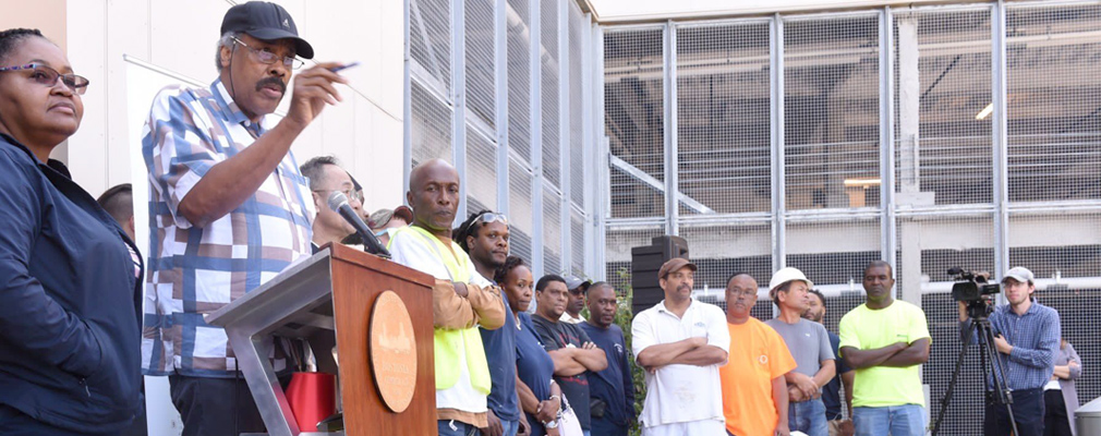 Photograph of contractors and construction workers participating in a ribbon-cutting ceremony, with one person speaking at a lectern.