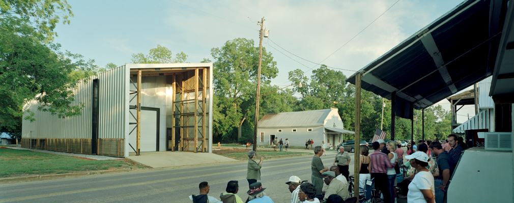 Photograph of a group of people seated along a road opposite a modern fire station.