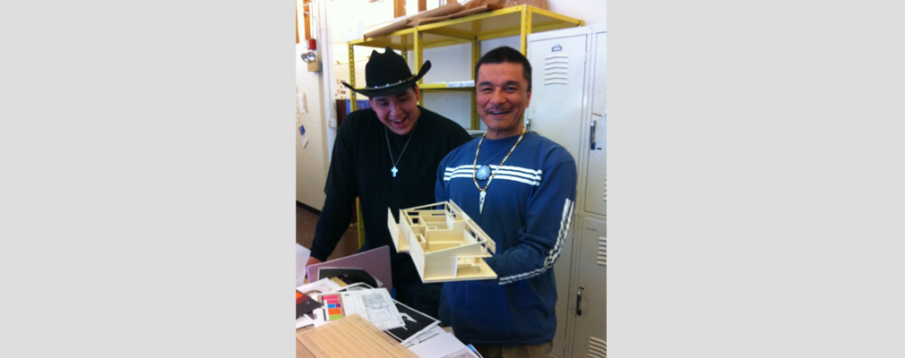 Photograph of two men, one of whom is holding a model house, in a classroom.