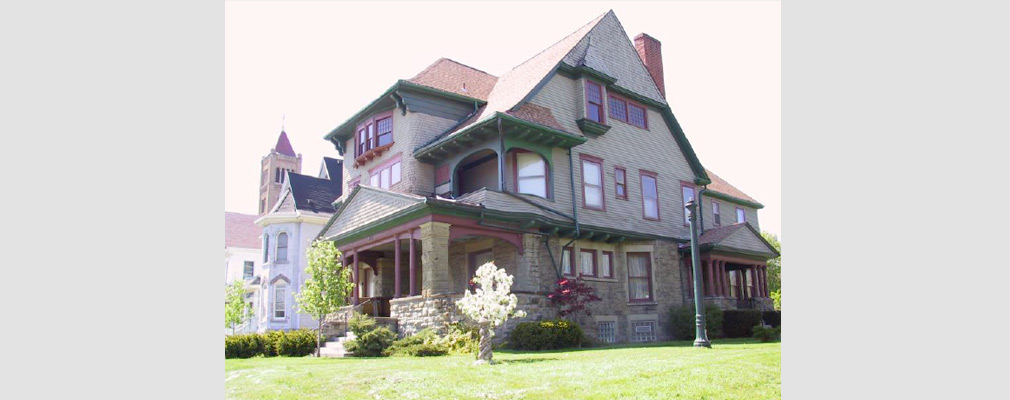 Photograph of two facades of a three-story Victorian style home with a yard in the foreground.