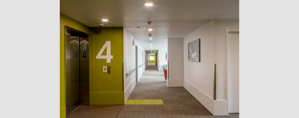 Photograph of a hallway with a handrail on one wall that leads to an elevator in the foreground.
