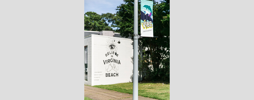 Photograph of a mural of words and pictograms saying “I believe in Virginia Beach,” with in the foreground, a lamp post and banner reading “ViBe Creative District.”