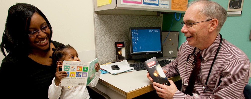 Photograph of a mother holding her daughter during a check-up with a doctor, who is holding a pamphlet titled, “Lead Poisoning.”