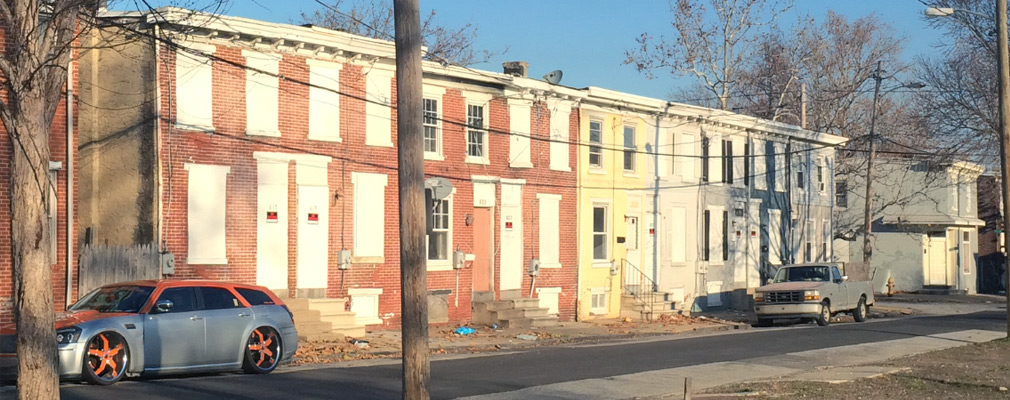 Photograph of nine vacant rowhouses, most of them with boarded windows. 