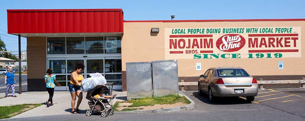 Photograph of the parking-lot side façade of a one-store grocery building, with a wall sign that says “Nojaim Bros. Market, since 1919.”