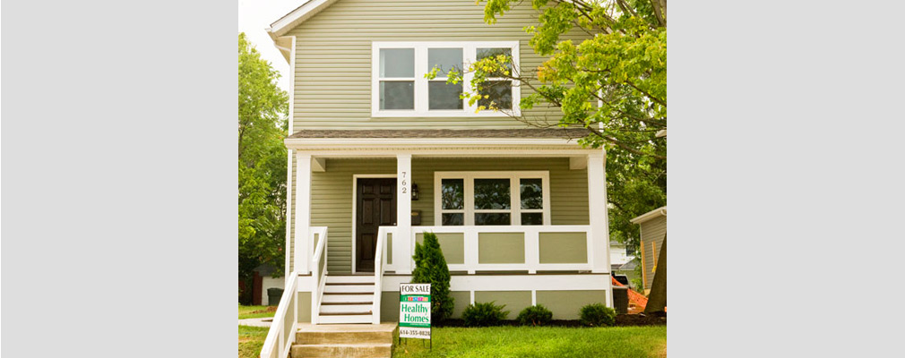 Photograph of the front façade of a two-story detached house, with a for sale sign in the front lawn that reads “Healthy Homes.”