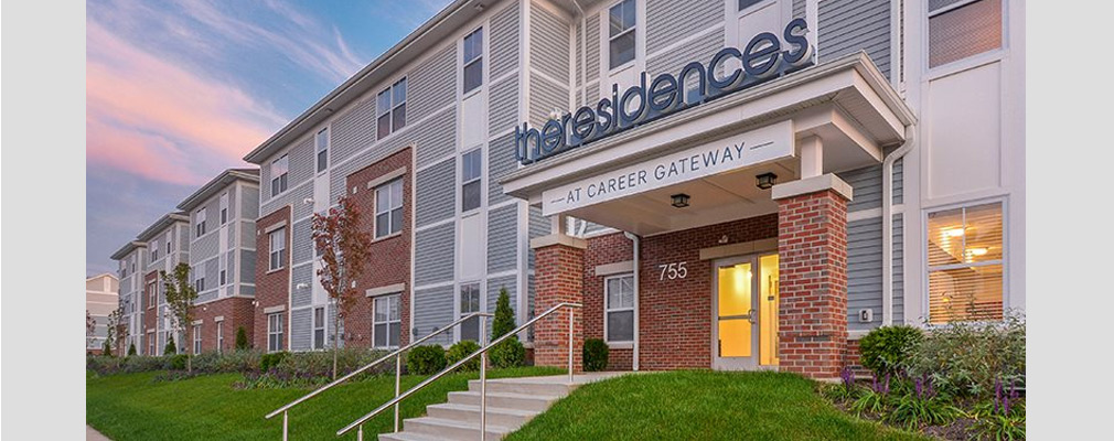 Photograph of the front façade of a three-story multifamily building with a sign over the entrance reading, “theresidences at career gateway.”