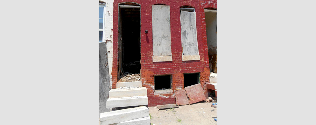 Photograph of the front of a blighted rowhouse with boarded-up windows, an empty doorframe, and debris in the doorway. 