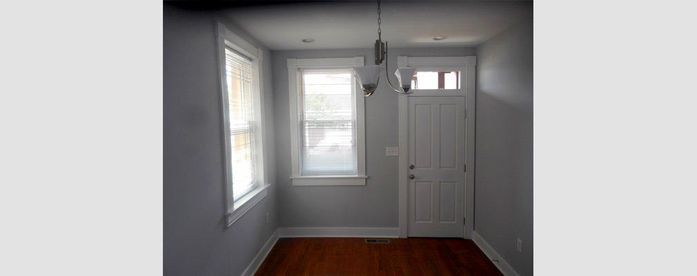 Photograph of a room in a rehabilitated rowhouse with wood flooring, wood-framed windows and a hanging light fixture. 