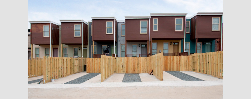 Photograph of the back of a group of rowhouses seen from an alley, with fenced-in paved parking spaces in the foreground. 