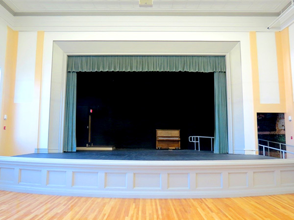 Photograph of a proscenium stage at one end of an auditorium with a wooden floor.