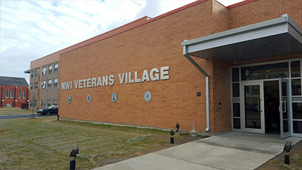 Photograph of the front door and façade of a one-story, flat-roofed building with a wall sign reading, “NWI Veterans Village”; a three-story residential wing is in the middle ground.
