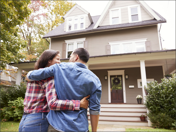 Two people embrace in front of single family home surrounded by trees.
