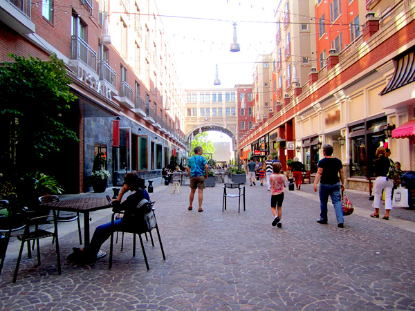 Several people walk along a pedestrian promenade and sit at outdoor tables between multistory, mixed-use buildings connected through an arched, enclosed walkway.