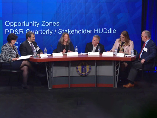 Moderator Erika Poethig, and panelists (left to right) Michael Kressig, Rachel Reilly, Frank Dickson, Fran Seegull, and Robert Stoker, sit at a table on stage in front of a banner stating “Opportunity Zones PD&R Quarterly/Stakeholder HUDdle.”