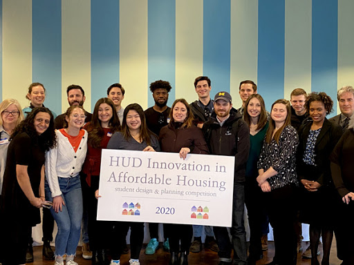 19 individuals, including students from the four finalist student teams associated with HUD’s Innovation in Affordable Housing Student Design & Planning Competition, stand together and hold a sign with the competition title.