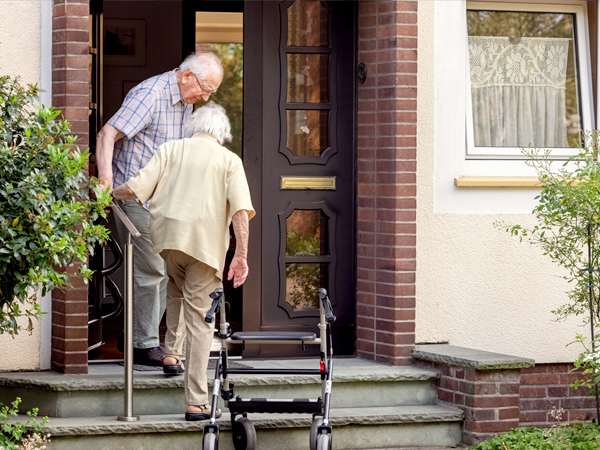 Two older adults walk up the steps in front of a house. A walker is visible in the foreground.