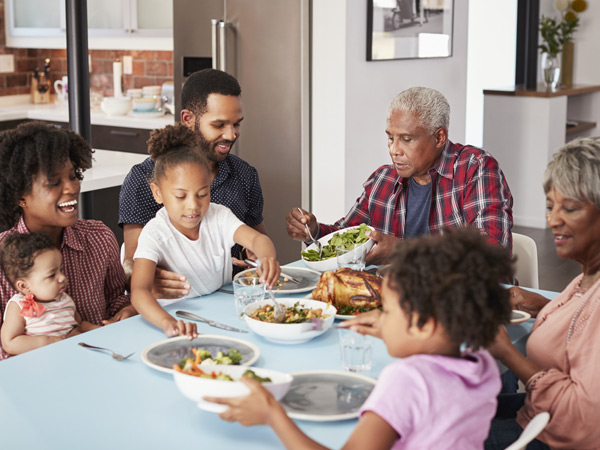 A multigenerational family, including two grandparents, two parents, and three children, are seated around a dinner table with food.